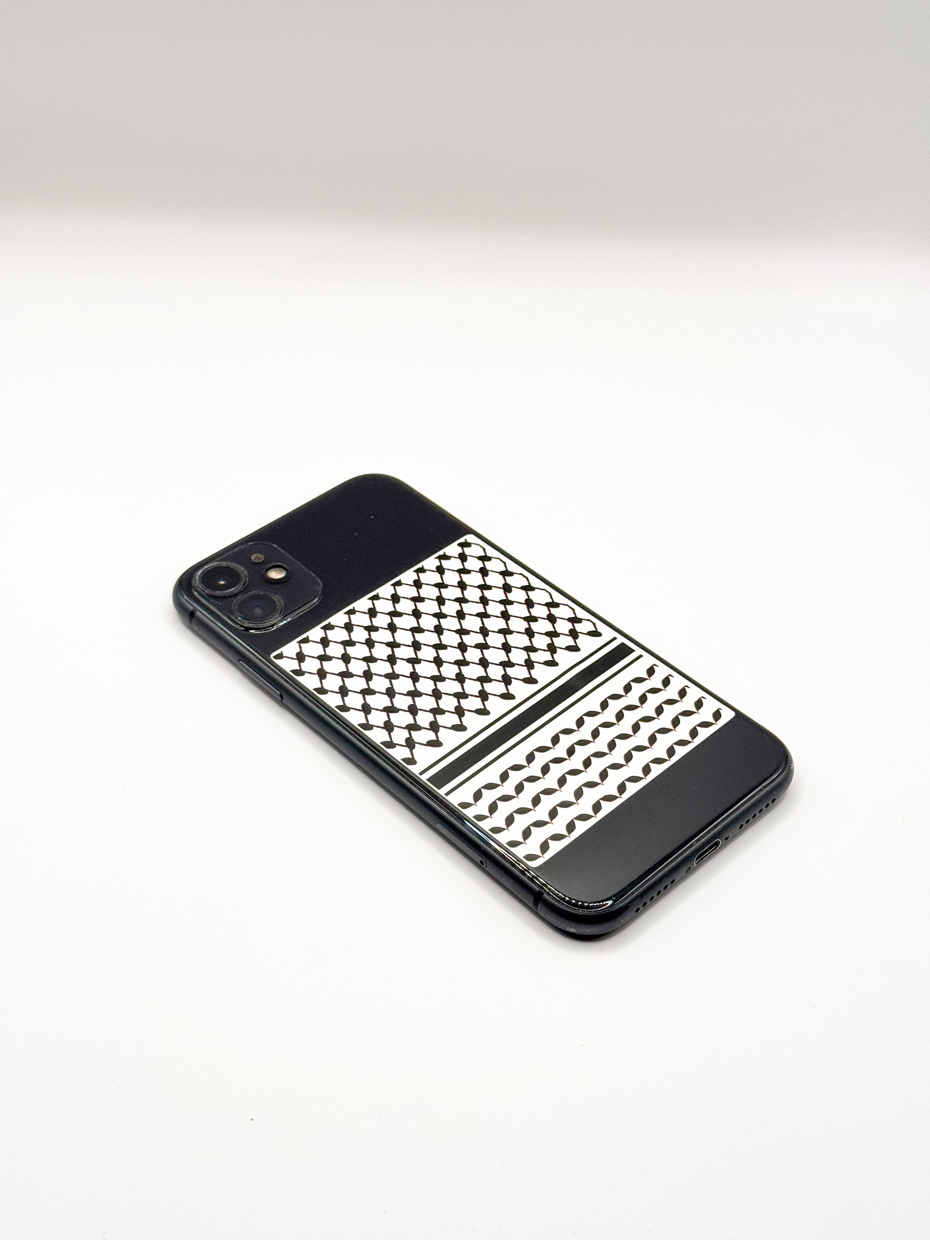 Mobiltelefon dekorert med kufiyeh-mønster, fotografert fra skrå vinkel mot lys bakgrunn.