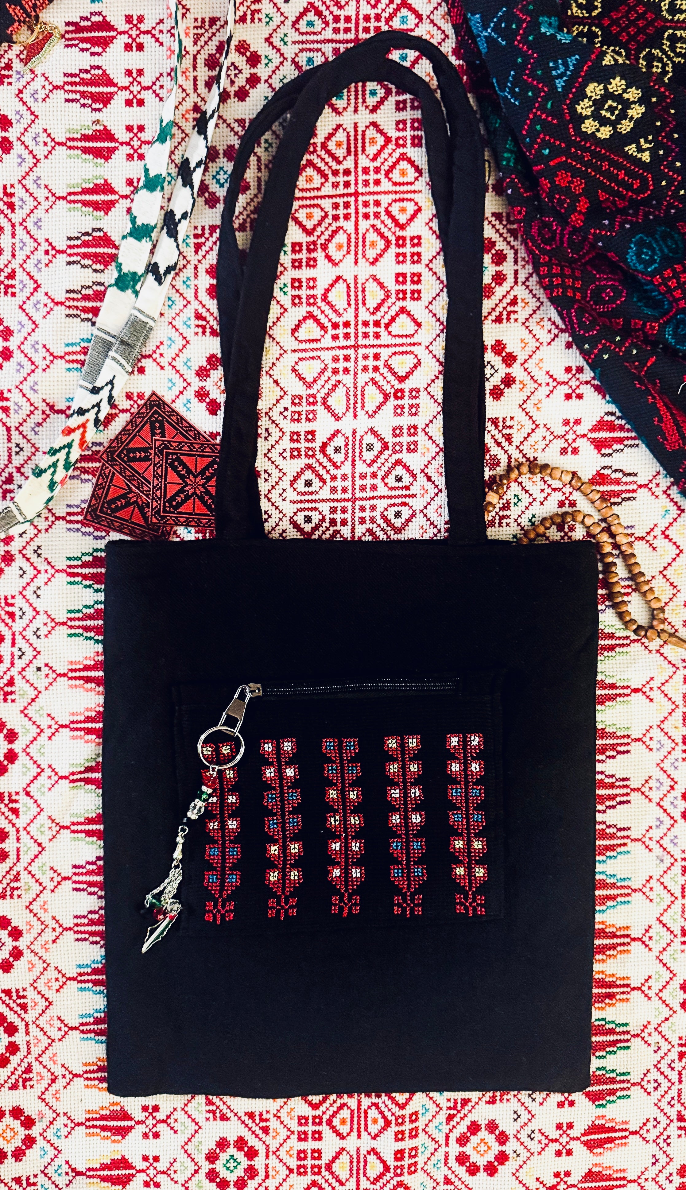 Sort tatreez tote bag i slitesterkt stoff med rødt palestinsk broderimønster, plassert på et tradisjonelt vevd tekstil med geometriske detaljer