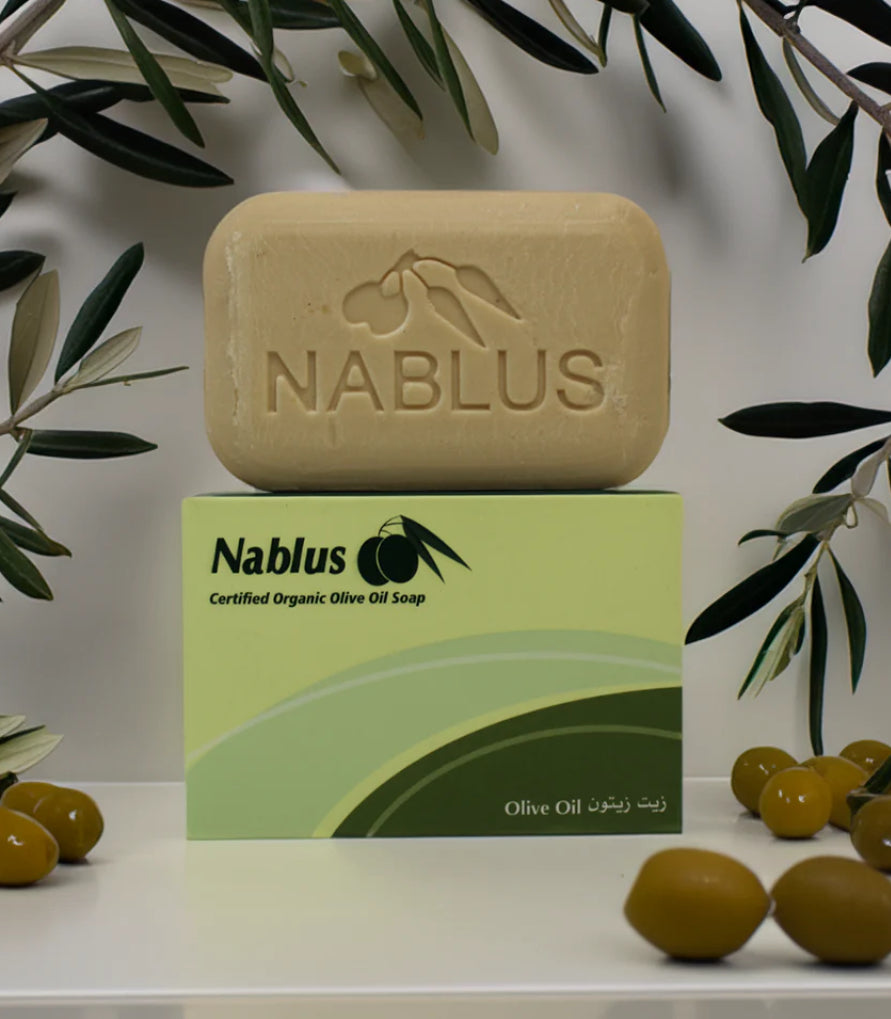 Håndlaget Nablus olivensåpe plassert på grønn eske, omgitt av oliven og olivengrener på hvit bakgrunn.