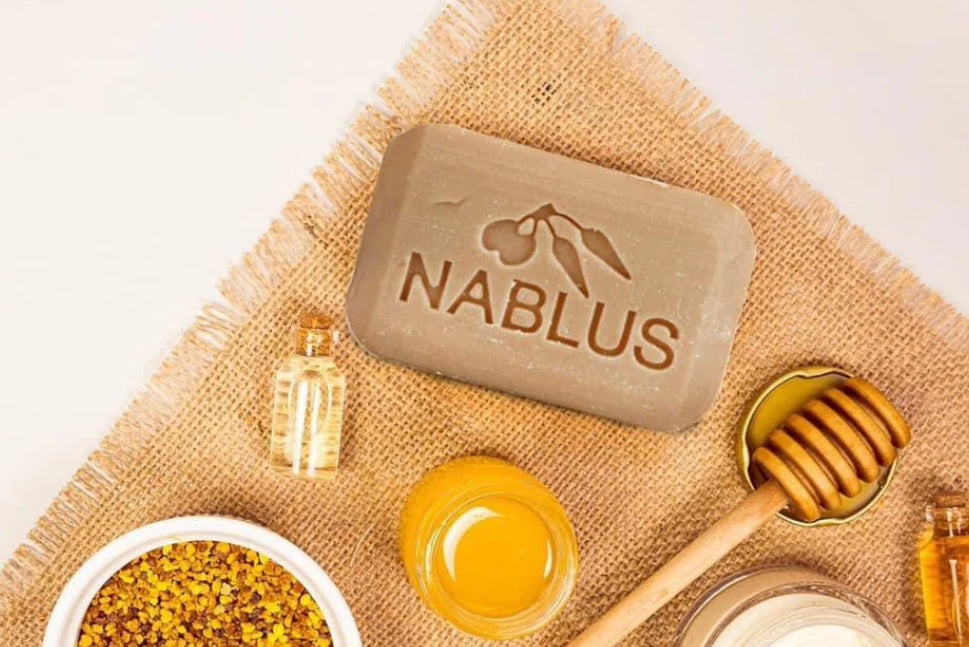 Nablus olivenoljesåpe med innpreget logo plassert på naturfarget lin, omgitt av honning, olje og naturlige ingredienser på lys bakgrunn.