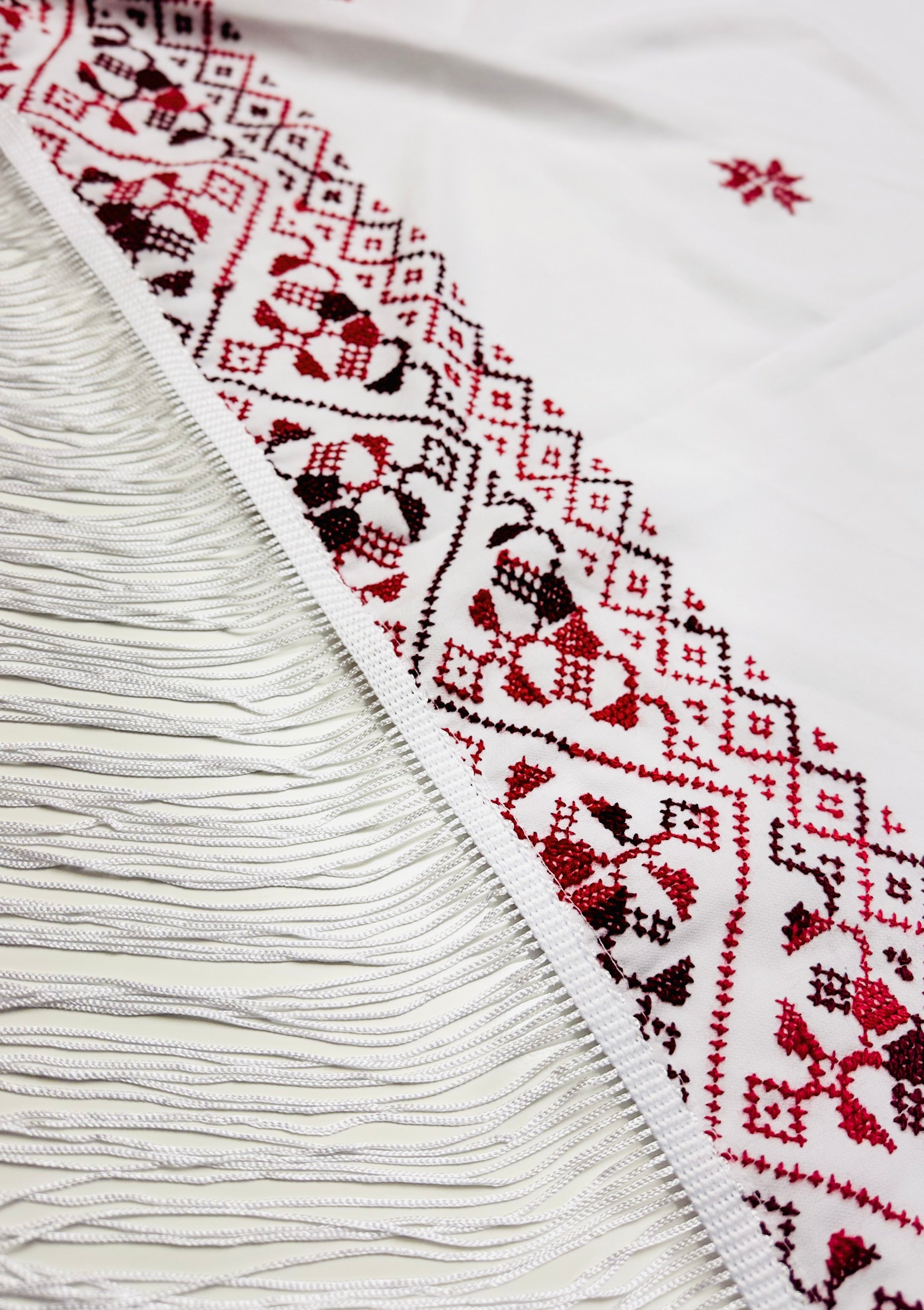 Nærbilde av hvitt tatreez-sjal med finere palestinsk broderimønster sett fra skrå vinkel, detaljert skjerf inspirert av tradisjonelt tatreez-håndverk